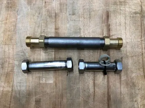 Drei Metallrohre mit verschiedenen Anschlüssen, darunter ein Danfoss-WMK-Passstück, auf einer Holzoberfläche angeordnet.